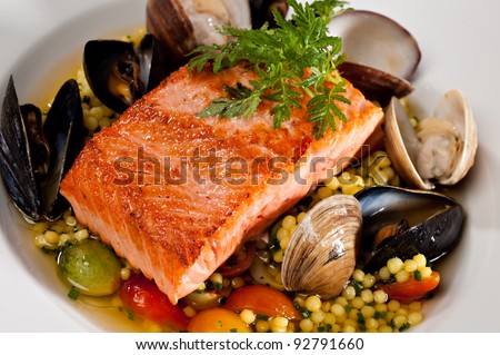 Beautifully plated salmon fillet garnished with colorful green parsley.  Served over a couscous and seafood salad featuring mussels, clams, tomatoes and brussels sprouts. Royalty-Free Stock Photo #92791660