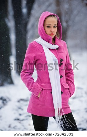Portrait of cute model in coat with hood and white scarf on the background of falling snowflakes in winter park