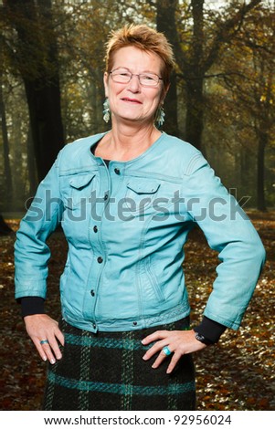 Senior woman short blond hair wearing glasses and blue jacket standing in autumn forest