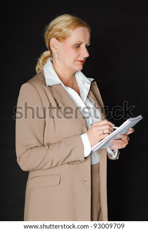 Studio portrait of senior blond woman in light brown suit and white shirt holding pen and paper isolated on black background