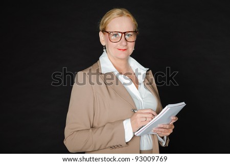 Studio portrait of senior blond woman wearing glasses in light brown suit and white shirt holding pen and paper isolated on black background