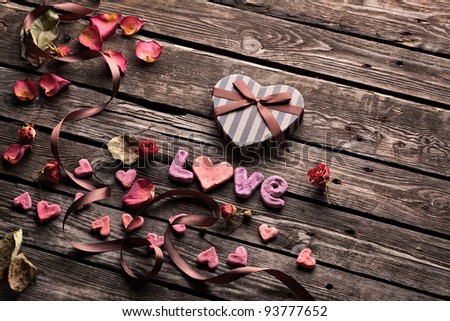 Word Love with Heart shaped Valentines Day gift box on old vintage wooden plates. Sweet holiday background with rose petals, small hearts, curved ribbon.