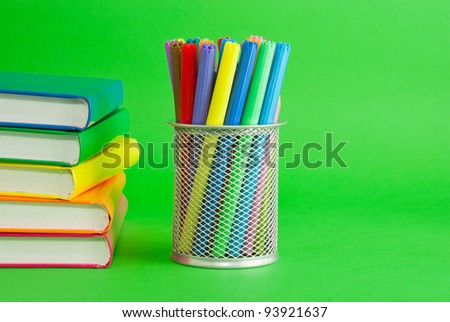 Stacks of colorful books and socket with felt pens against green background