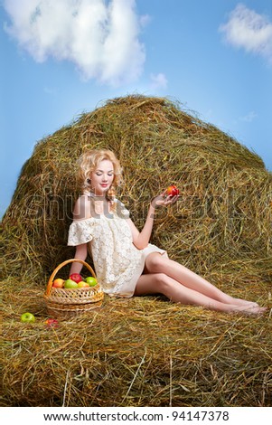 portrait of beautiful blonde country girl posing on yellow hay with basket of apples