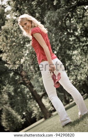 Smiling young woman holding her shoes and walking barefoot on grass in park