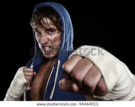 Boxer - street fighter in hoody fighting aggressive looking angry at camera hitting with hand wraps tape on hands. Young caucasian male fitness model isolated on black background.