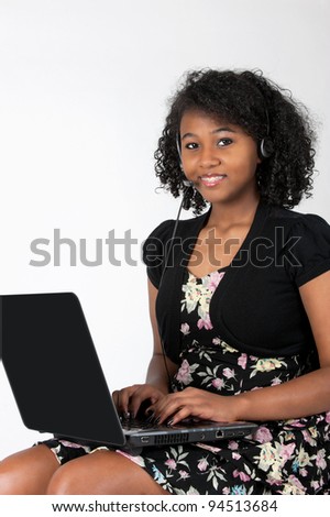 Beautiful Haitian African American teenage girl with braces on her teeth is wearing a headset and microphone and doing homework on a laptop computer with a big smile on her face.