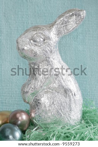 large silver rabbit with eggs sitting in artificial grass with blue background