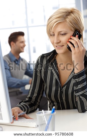 Cheerful businesswoman busy working, using desktop computer and speaking on mobile phone, multitasking.?