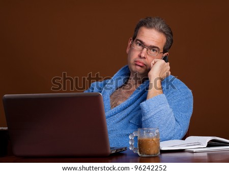 Middle aged man on the phone working at home in from of a laptop wearing a bathrobe and drinking a cup of coffee on a conference call
