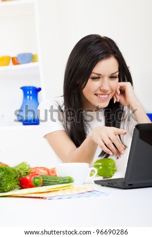 Beautiful young woman cooking looking at laptop screen with receipt in the kitchen, happy smile