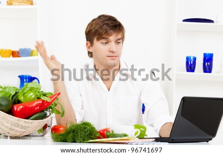 portrait of handsome young man cooking in the kitchen, problem prepare vegetable salad fail, looking at laptop screen with receipt for help with question hand gesture