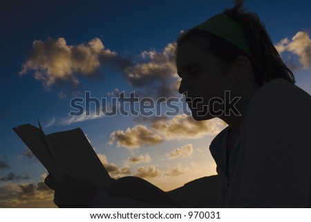 A young female silhouette reading a book in a beach. Backlighting with a sky as the background.
