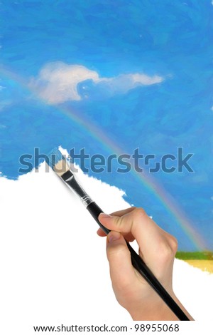 The hand with a brush draws a landscape.The blue sky, field and rainbow