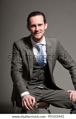 Smiling man brown long hair with expressive face wearing grey suit and blue tie. Isolated on grey background.