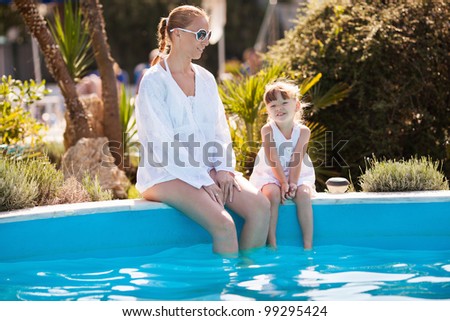 Happy mother and little daughter sitting on edge of the pool an playing