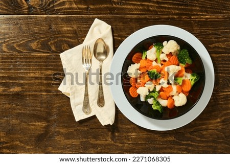 image from above of a black plate on a white one full of healthy vegetables on rustic brown wooden table and cutlery to the side on top of a white cloth napkin
