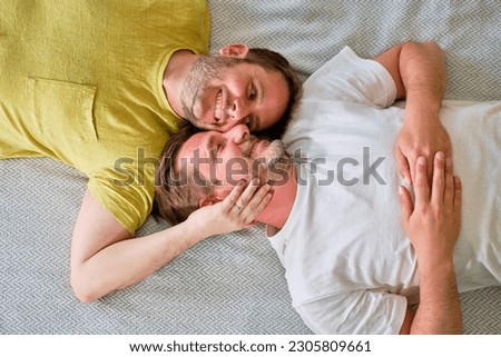 image from above of real gay couple over forty smiling lying on their backs in bed with their faces together