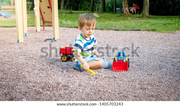 Image of adorable 3 years old toddler boy playing\
with sand and you truck and trailer in park. Child digging and\
building in sandpit