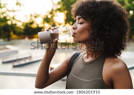 Image of african american sportswoman drinking water while working out on sports ground