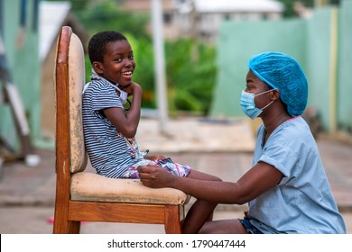 Image Of A An African Health Care Provider Wearing A Face Mask And Head Gear For Protection,squatting In Front Of A Little Girl Smiling And Looking Cheerful Outdoor