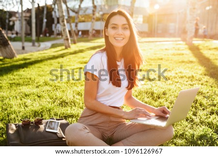 Image of amazing beautiful woman sitting on grass outdoors using laptop computer. Looking camera.