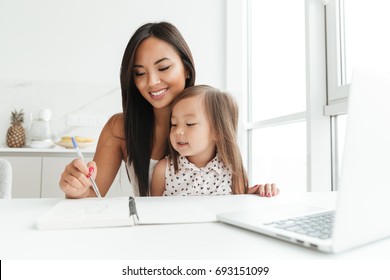 Image of amazing young mom sitting at the table with little cute asian girl at home indoors using laptop computer writing notes to notebook. Looking aside. స్టాక్ ఫోటో