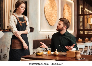 Image of angry bearded young man sitting in cafe. Looking at waiter.