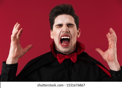 Image of angry vampire man with blood and fangs in black halloween costume screaming isolated over red wall