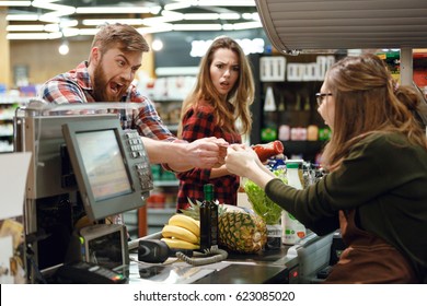 Image of angry young man standing in supermarket shop near cashier's desk doesn't give the credit card to cashier. Looking aside.