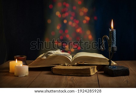 Image of an antique open book with bird feathers lying on a second closed book and a dark metal and wood candle holder with a blue candle on a wooden table with highlights on a dark background
