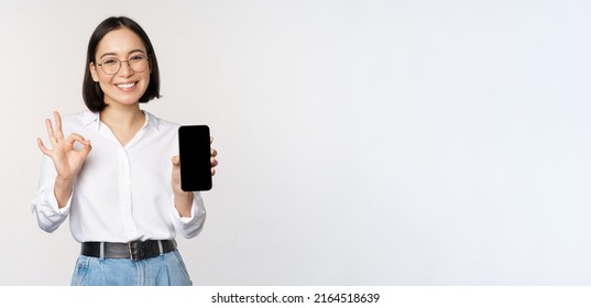 Image of asian businesswoman, showing smartphone screen, app interface and ok sign, recommending application on mobile phone, standing over white background - Powered by Shutterstock