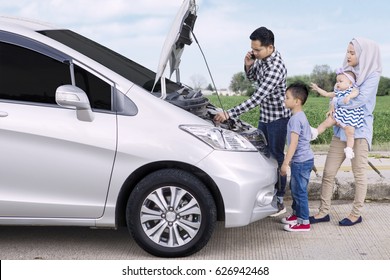 Image Of Asian Father Speaking On A Mobile Phone While Standing Front Of Broken Car With His Family On Side Of Road 