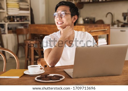 Image of asian happy man smiling and using laptop while sitting at table in cozy kitchen