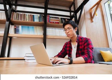 Image of asian male dressed in shirt in a cage and wearing glasses using laptop at the library. Looking at the laptop.