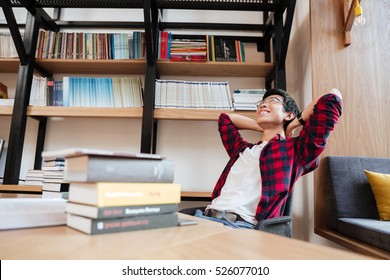 Image of asian man dressed in shirt in a cage and wearing glasses stretching near books and laptop at the library. - Powered by Shutterstock