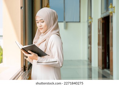 The image of an Asian Muslim woman in the Islamic religion in hijab in cream color. Standing reading the Qur'an and having a happy smiling face Staying in a beautiful mosque out of respect for God.