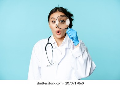 Image of asian woman doctor or nurse found smth, looking through magnifying glass and stare surprised, shocked, standing in uniform over blue background - Powered by Shutterstock