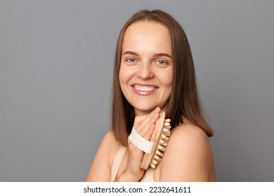Image of attractive beautiful woman uses dry body brush wants to have smooth clean skin, massaging her shoulder and arm, looking at camera with toothy smile, posing isolated over gray background.