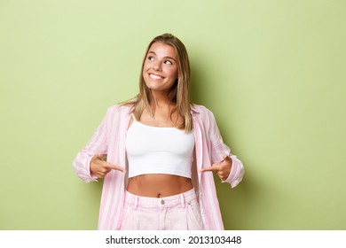 Image of attractive blond woman with flat belly, wearing stylish pink outfit, pointing fingers at stomach and looking at upper left corner with pleased smile