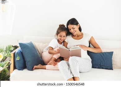 Image of attractive family pregnant woman and her little daughter smiling and reading book while sitting on sofa at home - Powered by Shutterstock