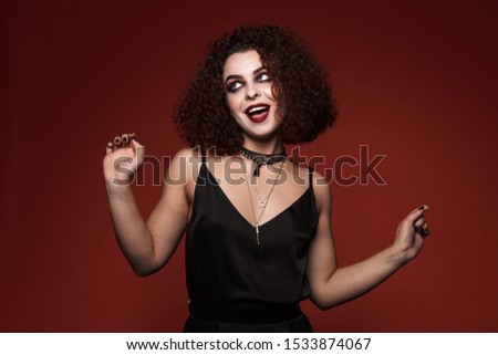 Image of attractive witch girl in black dress with halloween makeup smiling isolated over red wall