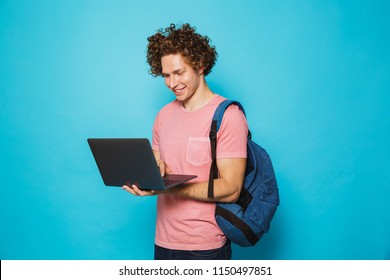 Image Of Attractive Young Guy With Curly Hair Wearing Casual Clothing And Backpack Holding And Using Open Laptop Isolated Over Blue Background