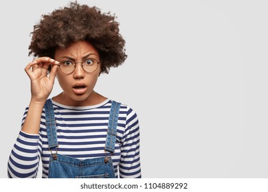 Image Of Beautiful African American Female Looks Scrupulously And Attentively Through Glasses, Tries To See Something In Darkness, Stands Against White Blank Wall With Copy Space For Your Promotion