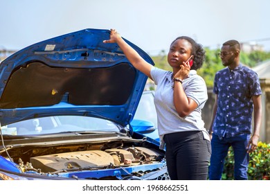 Image Of Beautiful African Lady With Smart Phone In Front Of An Opened Bonnet Of A Car, With Guy Bit Blurred At The Back- Transportation Concept