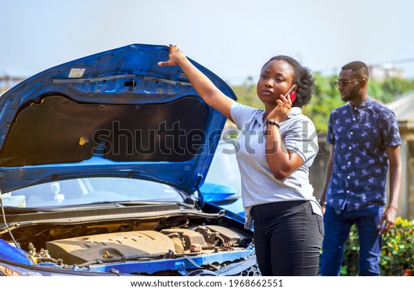 image of beautiful african lady with smart phone\
in front of an opened bonnet of a car, with guy bit blurred at the\
back- transportation\
concept