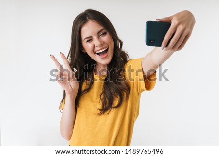 Image of beautiful brunette woman laughing and showing peace sign while taking selfie photo on cellphone isolated over white background