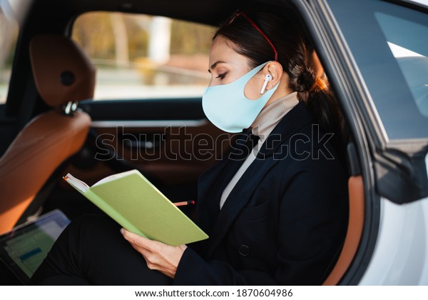 Image of beautiful business woman\
sitting in a car in face mask while holding\
notebook
