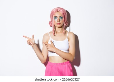 Image of beautiful caucasian girl in pink party wig and bright makeup pointing fingers left at your logo, showing banner about holidays or halloween, standing over white background