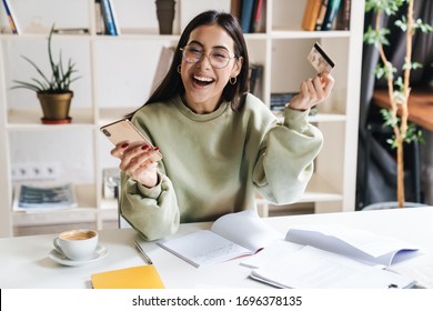 Image of a beautiful cheerful young girl student indoors studying using mobile phone holding credit card. - Powered by Shutterstock